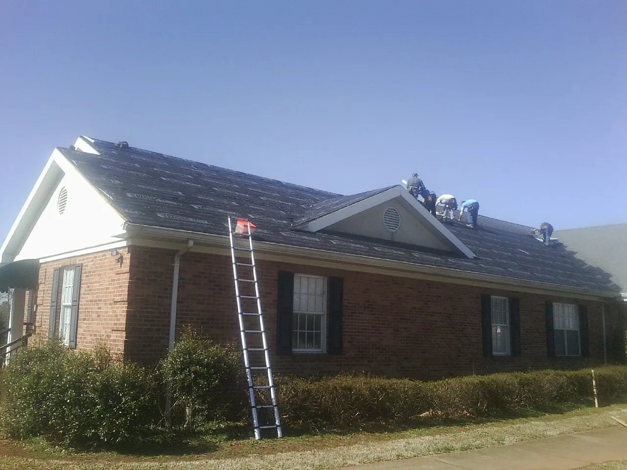 Crew tearing shingles off a brick ranch roof with a ladder set — Metal Roof Repair work by our crew in Twin City