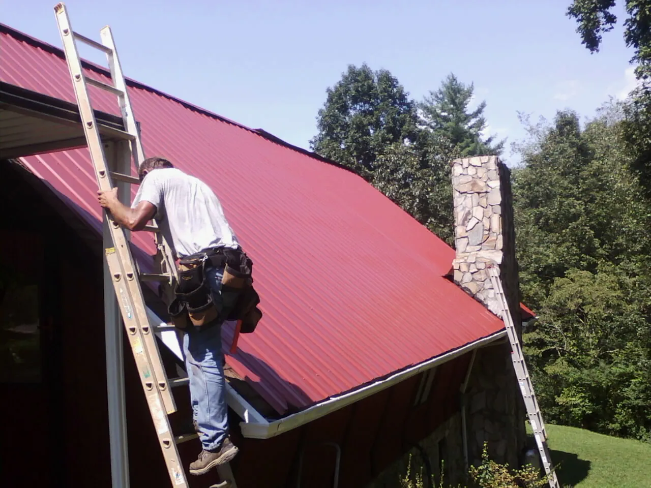Roofer on a ladder beside a red metal roof and stone chimney — Soffit & Fascia Repair by Roofing Brothers in Twin City